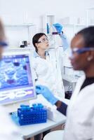 Serious thoughtful scientist analyzing color for liquid in glass flask. Multiethnic team of medical researchers working together in sterile lab wearing protection glasses and gloves. photo