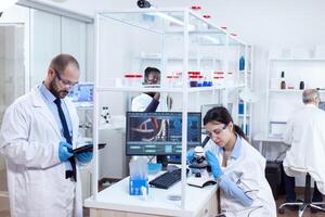 Chemist scientists doing vacine study in busy lab using tablet pc and miscroscope. Team of researchers doing pharmacology engineering in sterile laboratory for healthcare industry with african assistant in the background. photo