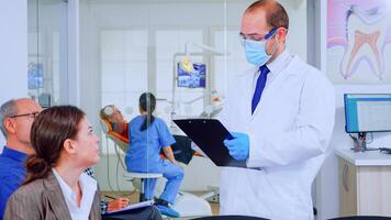 Doctor taking notes on clipboard about patient dental problems sitting on chair in waiting room of stomatological clinic. Assistant preparing old woman for examination in background photo