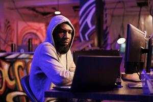 africano americano hacker codificación computadora virus a ataque base de datos. joven desarrollador en capucha agrietamiento contraseña y utilizando spyware a descanso dentro en línea servidor y robar información foto