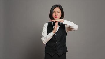 Asian receptionist expressing timeout symbol on camera, showing negativity and asking for a work break. Tired bored employee showing pause sign in studio over grey background. Camera A. photo