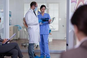 Dentist in stomatology waiting area holding patient x-ray explaining diagnosis to nurse in a crowded hallway with patient filling form. Dentistiry staff in reception. photo