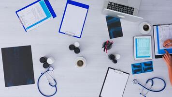 Top view of medical practitioner checking appointments using tablet, tacking notes on clipboard sitting on clinic desk with x-ray and medical equipment all around. Copy space, flat lay concept. photo