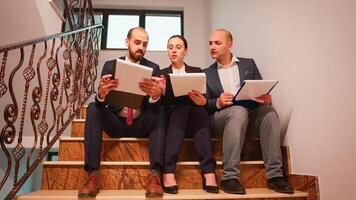 Project team of business workers looking on documents sitting on stairs explaining finance project doing overtime. Group of company manager and office executive on busy stairs of workplace. photo