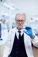 Experienced scientist in busy laboratory holding glass slide in microbiology laboratory. Senior researcher in sterile lab looking on microscope slide wearing lab coat. photo