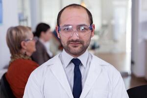 retrato de estomatólogo sonriente a cámara siendo en dental oficina mientras pacientes esperando él en antecedentes. odontología médico mirando en cámara web sentado en silla en estomatológico clínica. foto