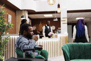 African American businessman drinking coffee while waiting for check-in procedure in hotel lobby. Tired black man traveler holding cup of espresso resting after long flight, waiting for room photo