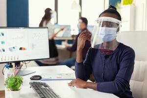 African manager in office with her coworkers wearing face mask against coroanvirus. Multiethnic business team working in financial company respecting social distance during global pandemic. photo