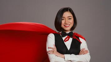 Hotel administrator wearing a red hero cloak on camera, feeling strong and powerful to help clients. Asian receptionist posing with confidence using a superhero cape over grey background. Camera B. photo