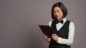 Asian hotel administrator browsing online website to check all reservations or room bookings, smiling and being confident. Receptionist using tablet in studio, hospitality industry. Camera B. photo