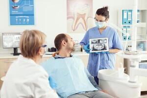Dentist woman in dental office discussing with sick man while nurse showing tooth x-ray image on tablet. Man patient waiting toothache treatment sitting on dental chair during medical examination photo