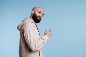 Smiling arab man inviting with hand and looking at camera with relaxed facial expression. Young bald bearded person asking to come over with arm gesture, posing for studio portrait photo