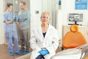 Senior stomatological woman looking into camera while sitting on dental chair in hospital stomatology room. Medical nurse discussing with patient during writing healdcare treatment photo