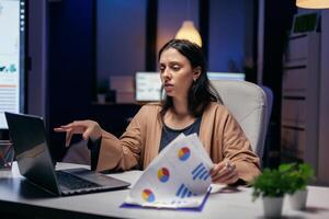 Tired employee reading article to help her finish deadline sitting at desk in empty office. Woman checking notes on clipboard while working on important project doing overtime. photo