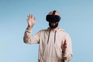 Young gamer enjoying metaverse with virtual reality headset. Man wearing vr helmet exploring entertainment activity in cyberspace while standing and making gesture with arms photo