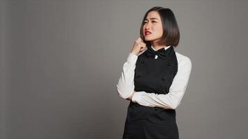 Restaurant hostess being thoughtful over grey background, thinking about new ideas to develop the menu for clients. Asian woman waitress feeling pensive, imagination in studio. Camera A. photo