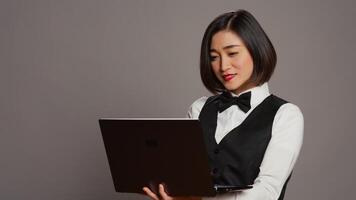 Asian hotel employee registering people using laptop at front desk, confirming online bookings and room reservations in studio. Receptionist posing with pc over grey background. Camera B. photo