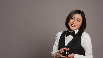 Woman hotel employee ringing front desk service bell in studio, working as a receptionist and registering guests in rooms. Asian concierge presenting a bell to call for assistance. Camera B. photo