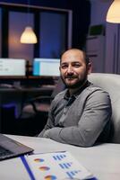 Focused freelancer smiling at camera resting hand on desk late at night. Smart businessman sitting at his workplace in the course of late night hours doing his job. photo