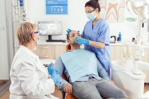 estomatólogo enfermero poniendo oxigeno máscara antes de diente cirugía sentado en dental silla en estomatología gabinete. mayor médico y enfermero trabajando en moderno ortodoncia oficina vistiendo máscara y guantes foto