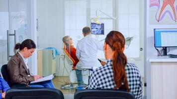 Patients sitting on chairs in waiting room of stomatological clinic filling in stomatological forms while doctor working in background. Concept of crowded professional orthodontist reception office. photo