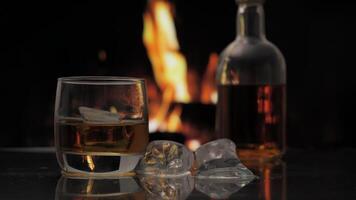 Glass and bottle of whiskey, scotch or bourbon stands on a table against background of a fireplace with a flame. Alcoholic drink and home comfort concept. video