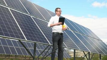 Engineer is looking at a solar panel and noting something in his tablet. Successful young businessman on the background of a solar power station video