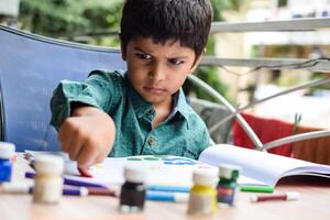 Smart Indian little boy perform thumb painting with different colourful water colour kit during the summer vacations, Cute Indian Kid doing colourful thumb painting drawing on wooden table photo