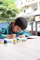 Smart Indian little boy perform thumb painting with different colourful water colour kit during the summer vacations, Cute Indian Kid doing colourful thumb painting drawing on wooden table photo