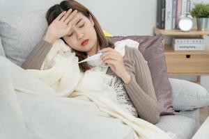 joven asiático mujer teniendo alto fiebre mientras comprobación cuerpo temperatura, hembra estornudos y líquido nariz con estacional influenza, alérgico, digital termómetro, virus, coronavirus, enfermedad, respiratorio foto