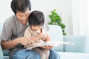 Happy Asian father relax and read book with baby time together at home. parent sit on sofa with daughter and reading a story. learn development, childcare, laughing, education, storytelling, practice. photo