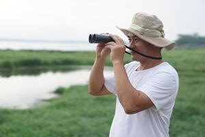 Handsome Asian man ecologist is surveying nature at the lake, hold binoculars. Concept, nature exploration. Ecology study. Pastime activity, lifestyle. Explore environment photo