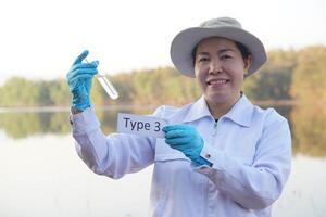 asiático mujer ecologista sostener tubo de muestra agua y etiqueta con palabra tipo 3 para inspeccionar y investigación agua calidad a el lago. concepto, explorar, análisis agua calidad desde natural fuente. ecología. foto