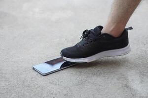 Closeup man wear black shoe, step on smartphone on ground, cause it crack. Concept, damage with things in daily life from accident or careless. Mobile phone warranty claim for broken photo