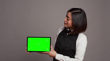 Woman waitress holding tablet with greenscreen display on camera, presenting blank mockup template in studio. Asian restaurant employee showing device running chromakey screen. Camera B. video