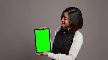 Asian restaurant hostess showing greenscreen display in studio, presenting isolated mockup layout on tablet. Waitress working with blank copyspace chromakey template on screen. Camera B. video