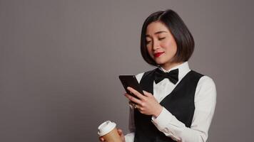 Concierge personnel drinking cup of coffee in studio, browsing smartphone social media apps. Asian receptionist enjoying caffeine refreshment while she uses mobile phone. Camera B. video