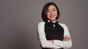 Restaurant hostess posing with confidence in studio, standing with arms crossed over grey background. Asian waitress barista with gourmet serving expertise smiling on camera. Camera B. video