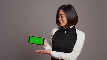 Catering employee holding phone with greenscreen display, pointing at chromakey design on smartphone. Asian waitress using isolated copyspace template over grey background in studio. Camera B. video