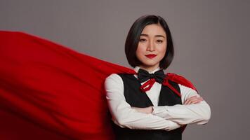 Asian receptionist posing with red super hero cape on camera, feeling confident and powerful while she wears a cloak as a hero. Hotel concierge operator standing with arms crossed. Camera B. video