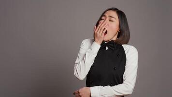 Exhausted waitress with apron yawning over grey background, feeling overworked and dealing with burnout after multiple restaurant events. Asian catering worker feeling tired in studio. Camera B. video