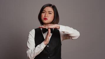 Asian receptionist expressing timeout symbol on camera, showing negativity and asking for a work break. Tired bored employee showing pause sign in studio over grey background. Camera B. video