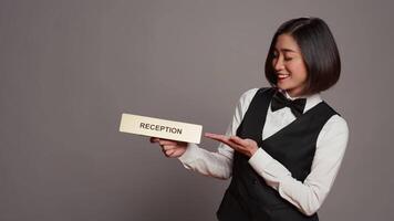 Hotel concierge holding sign to indicate direction for reception desk, presenting indicator to help guests and offer assistance in studio. Employee with uniform and bow uses wall pointer. Camera B. video