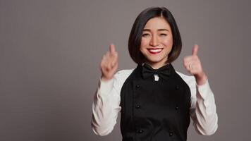 Smiling waitress giving thumbs up sign in front of camera, working in a five star restaurant to serve tables. Asian server with apron showing approval with okay symbol in studio, agreement. Camera B. video