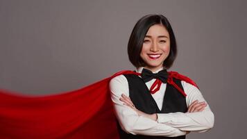 Hotel administrator wearing a red hero cloak on camera, feeling strong and powerful to help clients. Asian receptionist posing with confidence using a super hero cape over grey background. Camera B. video