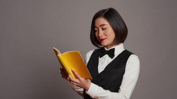 Asian receptionist reading a literature book over grey background, enjoying lecture hobby with a novel story in studio. Hotel concierge employee reads a fiction tale for knowledge. Camera B. video