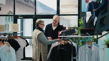 ouderen klanten analyseren elegant overhemden Aan hangers Bij kleinhandel op te slaan, op zoek Bij nieuw kleding lijn Aan Scherm Bij boetiek. senior paar zoeken voor formeel dragen, chique levensstijl. camera b. video