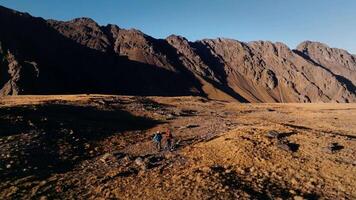 Two men with backpacks walk along a plateau high in the mountains against a backdrop of snow-capped mountains and high rocky walls. Aerial view video