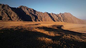 Two men with backpacks are trekking along a mountain plateau high in the mountains surrounded by rocky mountains in late autumn. Aerial view following travelers video