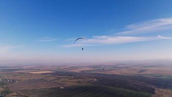 aérien voir, stabilisé métrage le long de le horizon. une Masculin parapente mouches entouré par des champs et collines. en retard automne. bleu blanc parapente aile dans ensoleillé temps video